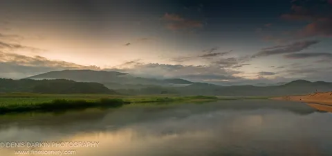 Calm delta waters at Tachingouza Bay reflect a sky gradient from amber to slate blue, with green marshland on the left bank and hazy mountains beyond.