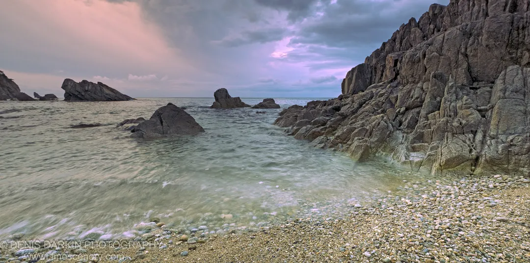 Steep craggy cliff with vertical striations rising above a pebble shore at the Far East Marine Reserve, Primorsky Krai, under a purple-tinted sky.