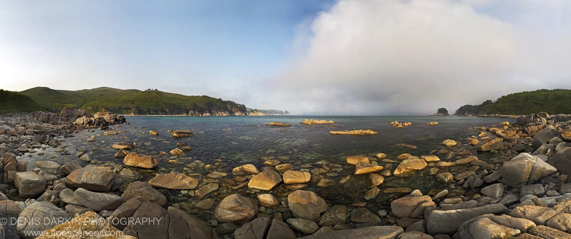 Smooth, sun-warmed boulders line the shallow bay foreground, with rolling green hills on either side of the cove and a cloud bank to the right.