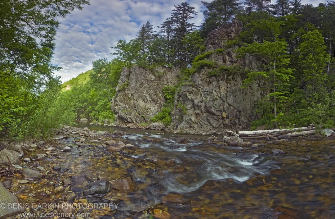 A clear stream flows over smooth multi-toned stones at the base of a steep rocky cliff in Sikhote Alin, Primorsky Krai.