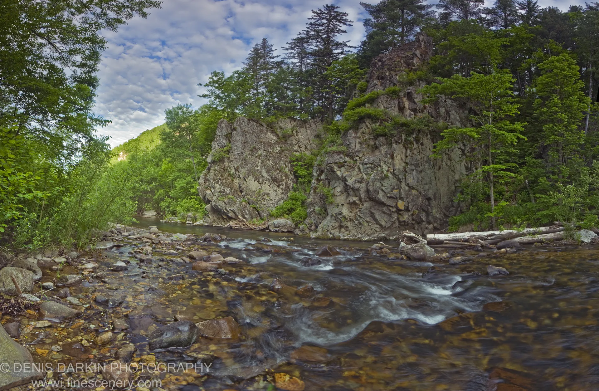 A clear stream flows over smooth multi-toned stones at the base of a steep rocky cliff in Sikhote Alin, Primorsky Krai.