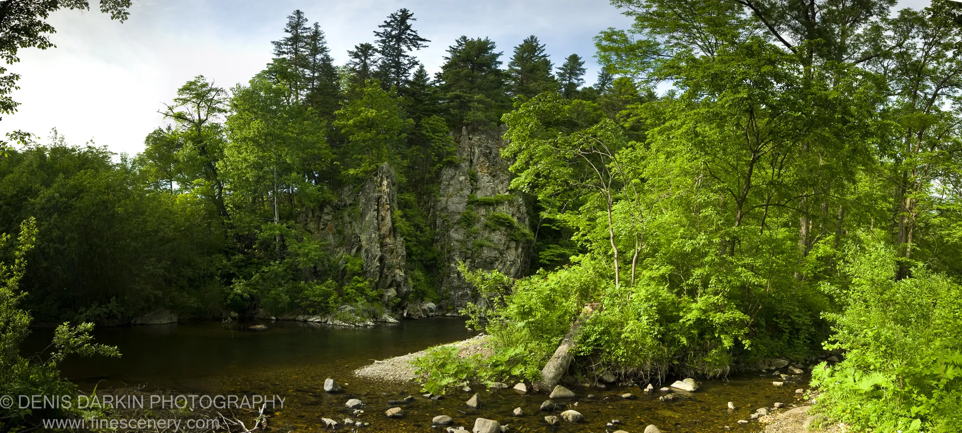 Lush deciduous foliage and moss-covered cliffs above the shallow, stone-dotted waters of a forest creek in Sikhote Alin.