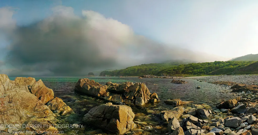 Sunlit granite boulders emerge from crystal-clear water in the foreground as a dense cloud bank rolls over the green hills beyond.