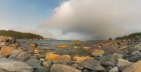 Smooth boulders in shallow turquoise water lead to green headlands beneath a billowing cloud formation with a faint rainbow arc.