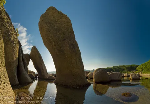 Massive leaning granite monoliths rise from shallow tidal pools at Langou Bay, their reflections mirrored in still water under a deep blue sky.