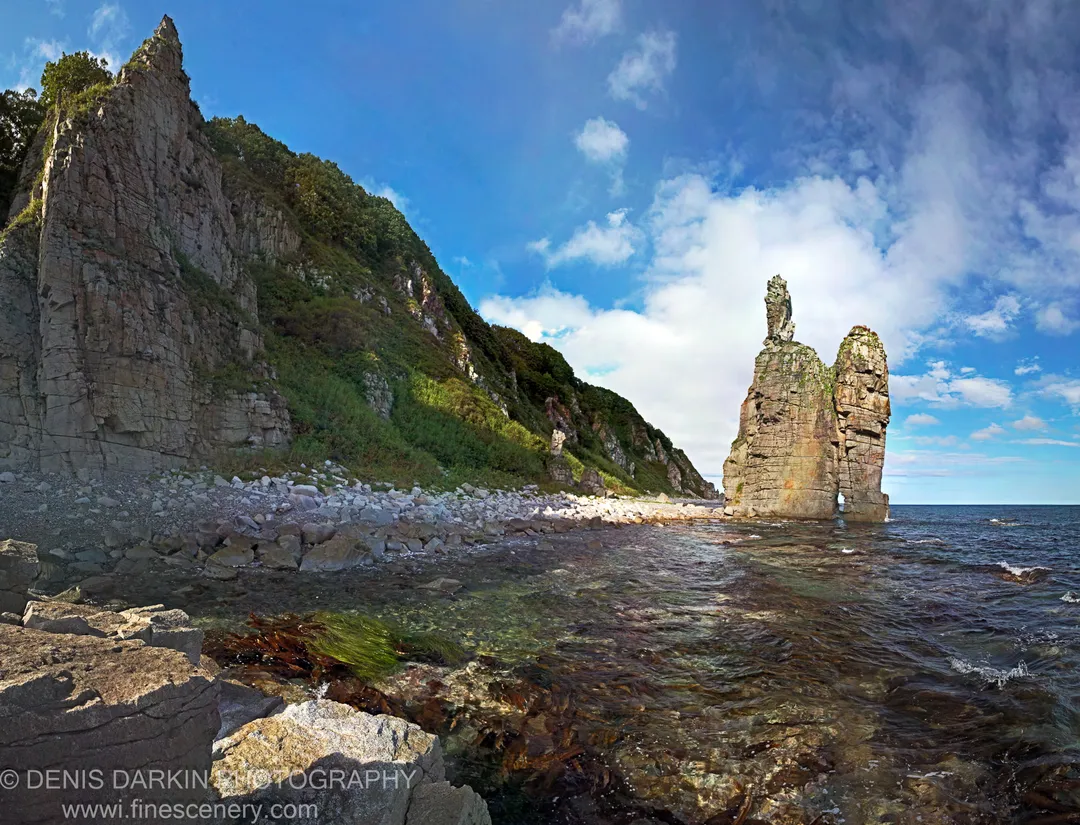 Isolated sea stack rising from clear, shallow coastal water near Dalnegorsk, with submerged seaweed and stones visible through the transparent surface.