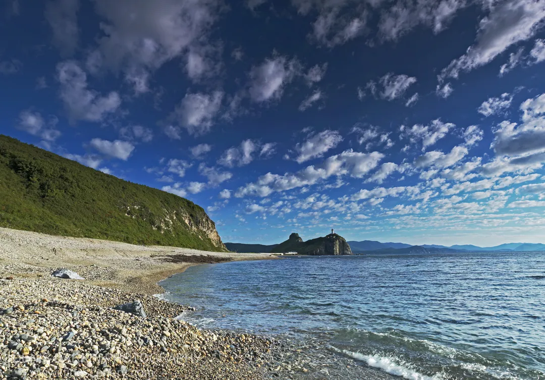 Pebble beach and deep blue water at Dalnegorsk with distant rocky headlands and a lighthouse visible under a clouded sky.