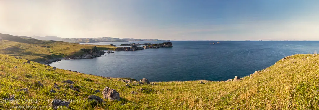 Sunlit grass and dark stones slope toward rocky headlands curving around a sheltered bay, with distant islands on the horizon, Far East Marine Reserve.