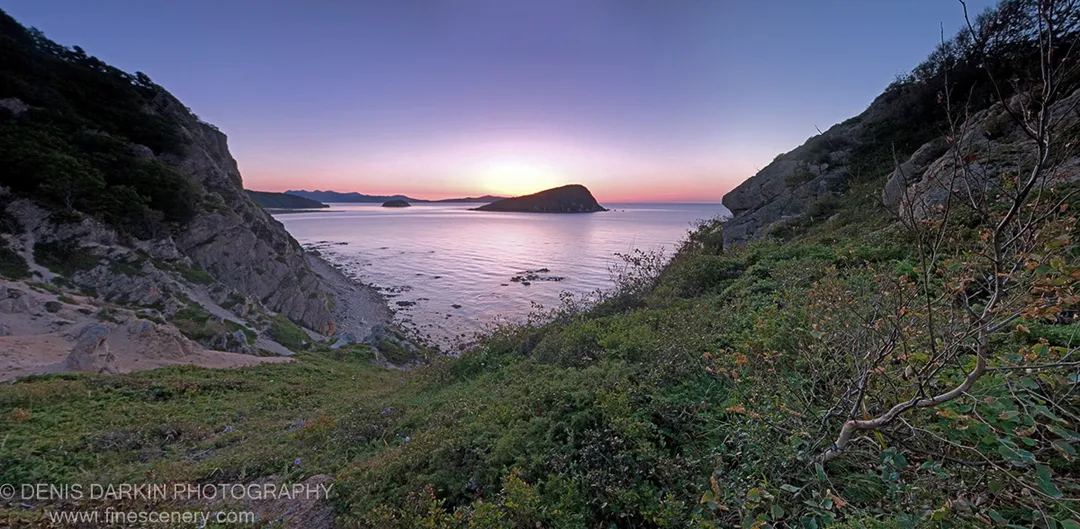 Panoramic view from rocky, vegetation-covered slopes of Petrova Island at sunrise, with distant islands silhouetted against a soft morning sky over calm water.