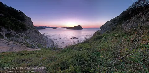Panoramic view from rocky, vegetation-covered slopes of Petrova Island at sunrise, with distant islands silhouetted against a soft morning sky over calm water.