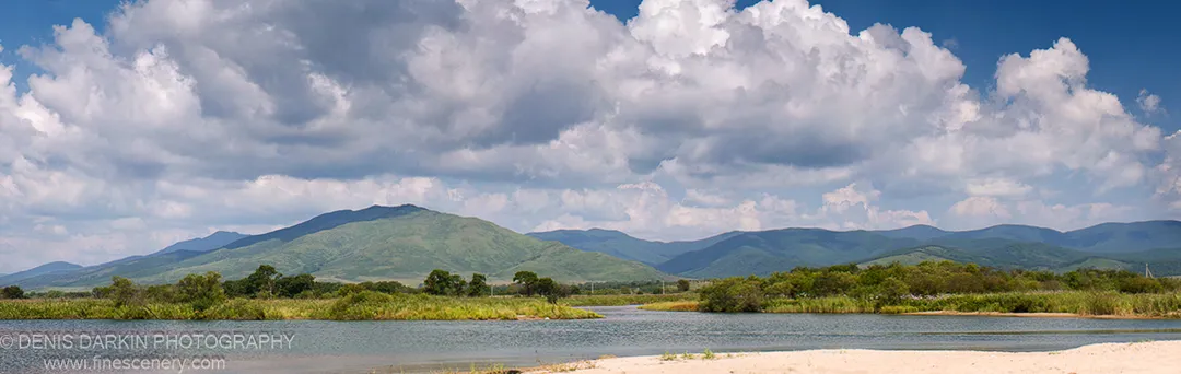 Wide river meets a sandy shore below towering cumulus clouds, with green rolling mountains behind, Narva Bay, September 2007.