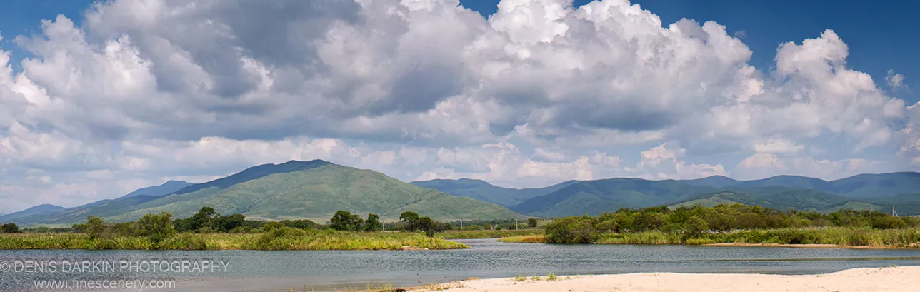 Wide river meets a sandy shore below towering cumulus clouds, with green rolling mountains behind, Narva Bay, September 2007.
