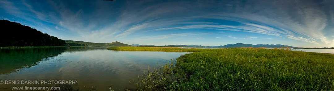 A sunlit grassy spit curves into calm water with a dark hill silhouetted on the opposite bank, Narva Bay, September 2007.