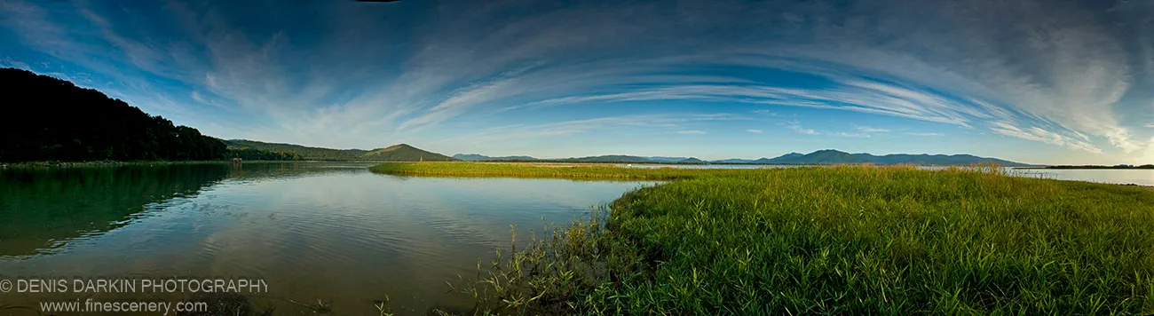 A sunlit grassy spit curves into calm water with a dark hill silhouetted on the opposite bank, Narva Bay, September 2007.