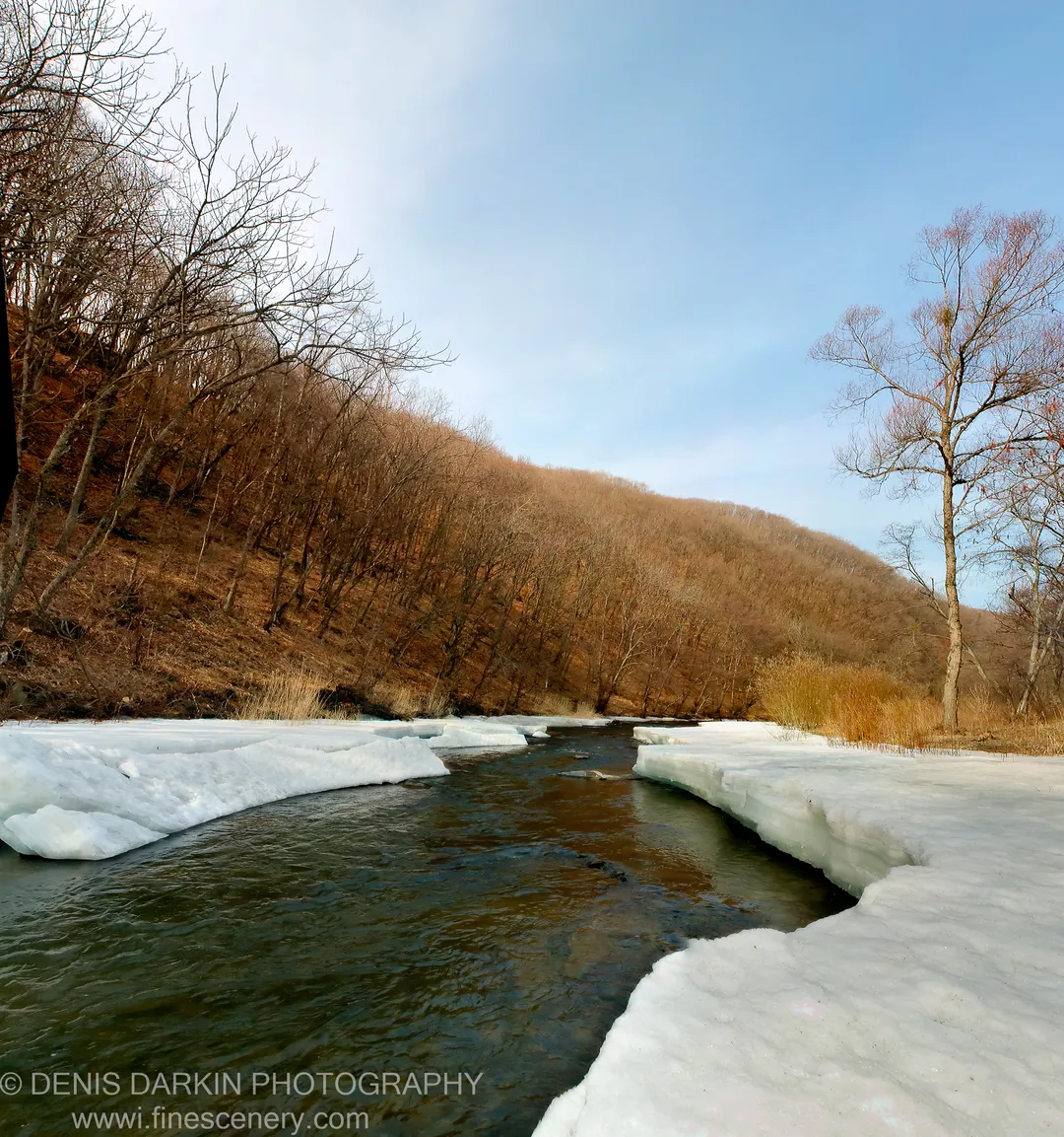 A river flows through a steep valley in Primorsky Krai, thick ice sheets on the banks and bare trees rising on either side.