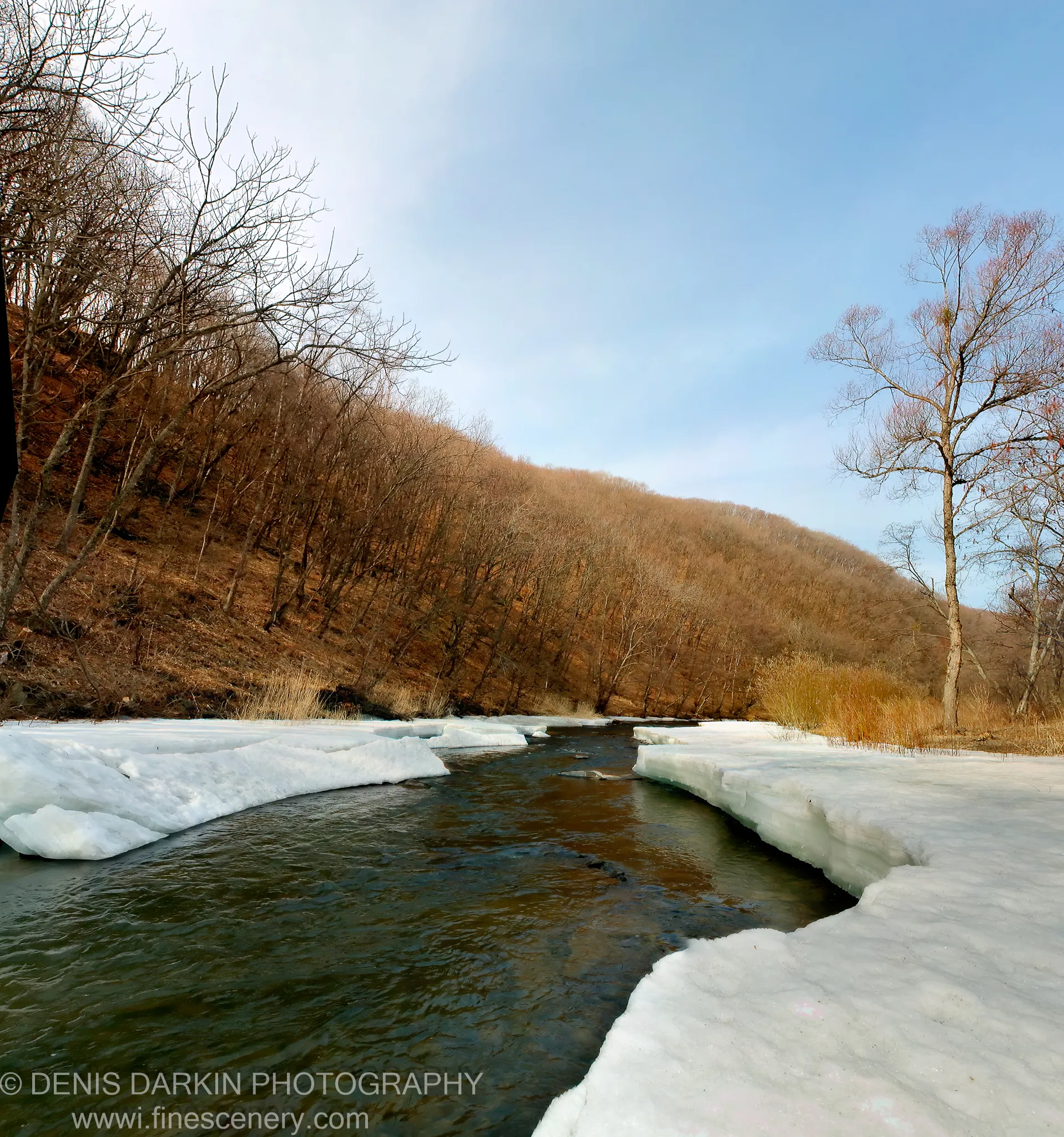 A river flows through a steep valley in Primorsky Krai, thick ice sheets on the banks and bare trees rising on either side.