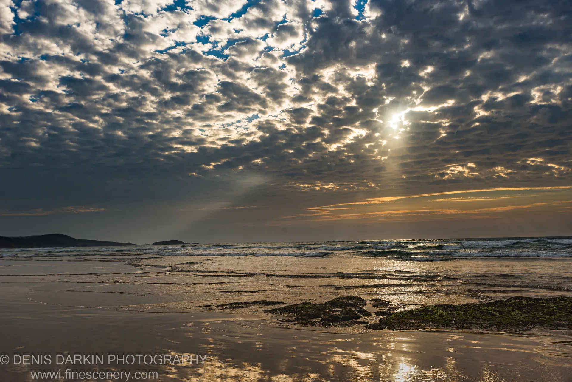 Crepuscular rays spread across the horizon at One Mile Beach at dawn, with dark seaweed-covered rocks and wet sand ripples below.