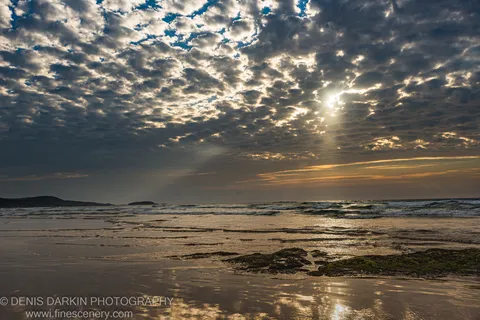 Crepuscular rays spread across the horizon at One Mile Beach at dawn, with dark seaweed-covered rocks and wet sand ripples below.