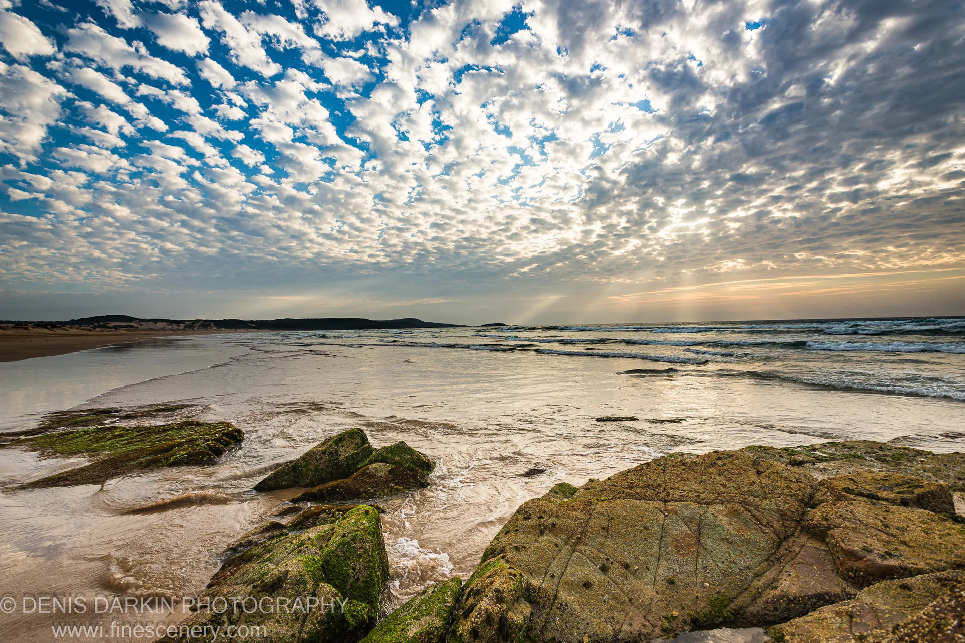 Morning sunbeams break through altocumulus cloud at One Mile Beach, Port Stephens, casting warm light over moss-covered boulders and rolling surf.