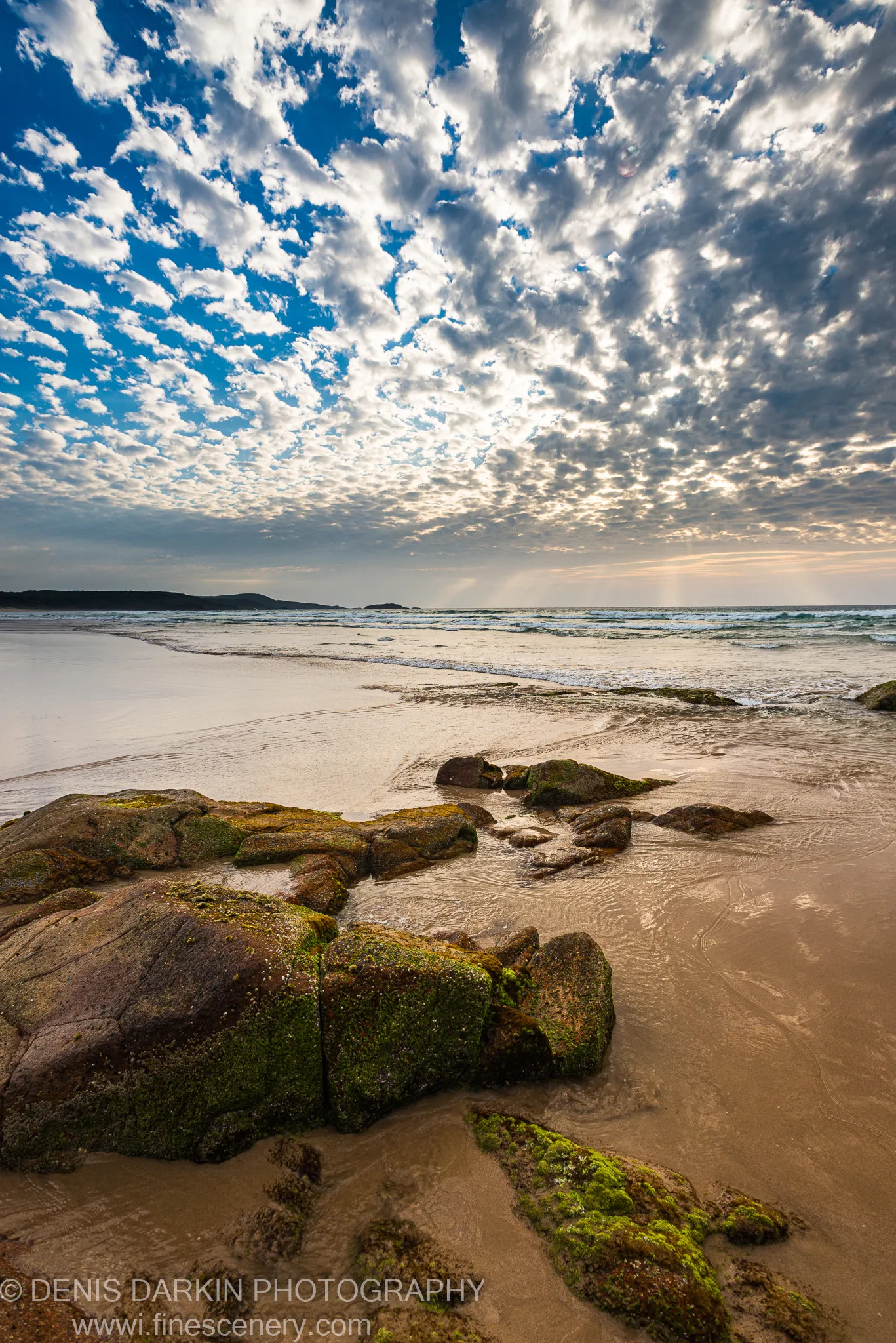 Moss-covered boulders on the wet sand at One Mile Beach as the sun rises above a textured cloud ceiling in early December.