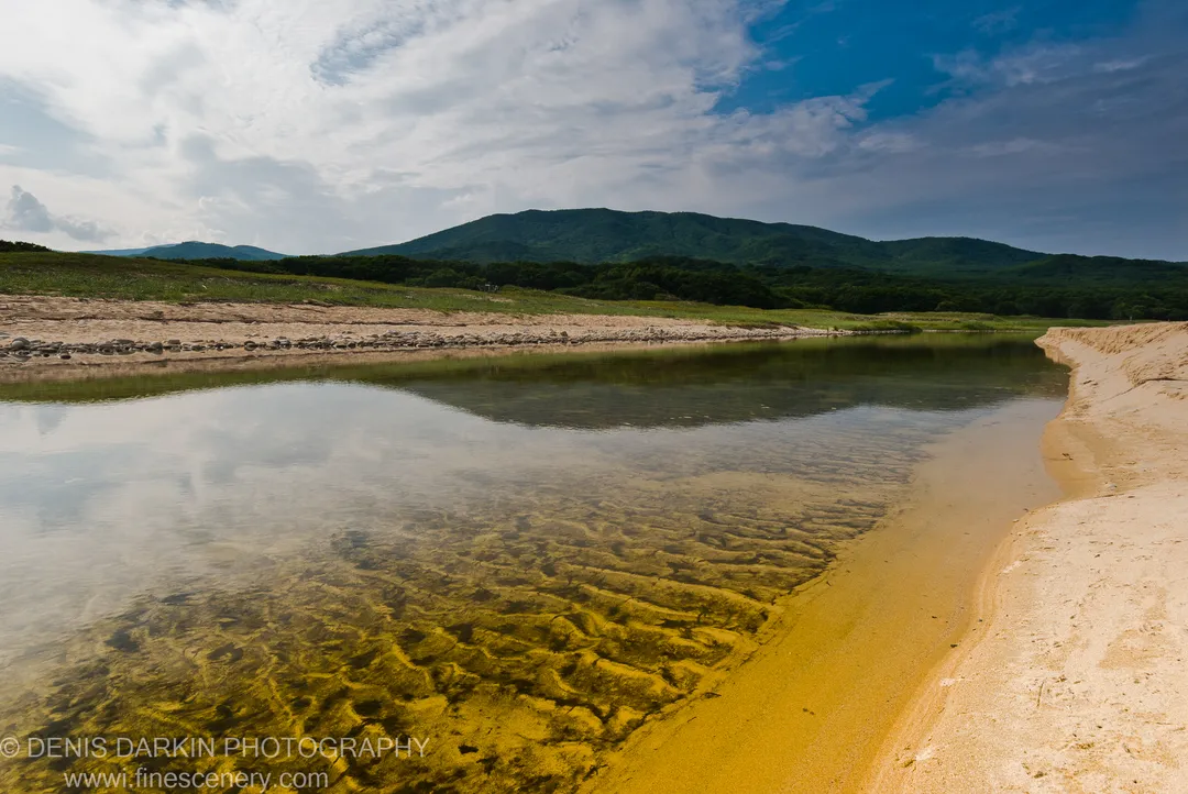 A shallow sandy bay with submerged ripple patterns in the foreground and dense forested hills rising under a scattered cloud sky, Lazovsky Reserve.