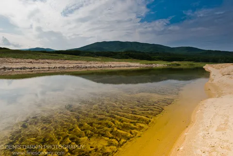 A shallow sandy bay with submerged ripple patterns in the foreground and dense forested hills rising under a scattered cloud sky, Lazovsky Reserve.