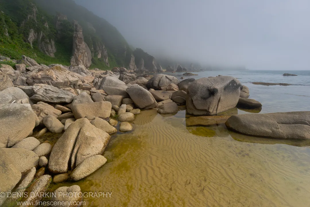 Weathered boulders rise from shallow water along the Lazovsky Reserve coast, with a steep green cliff and misty horizon beyond.
