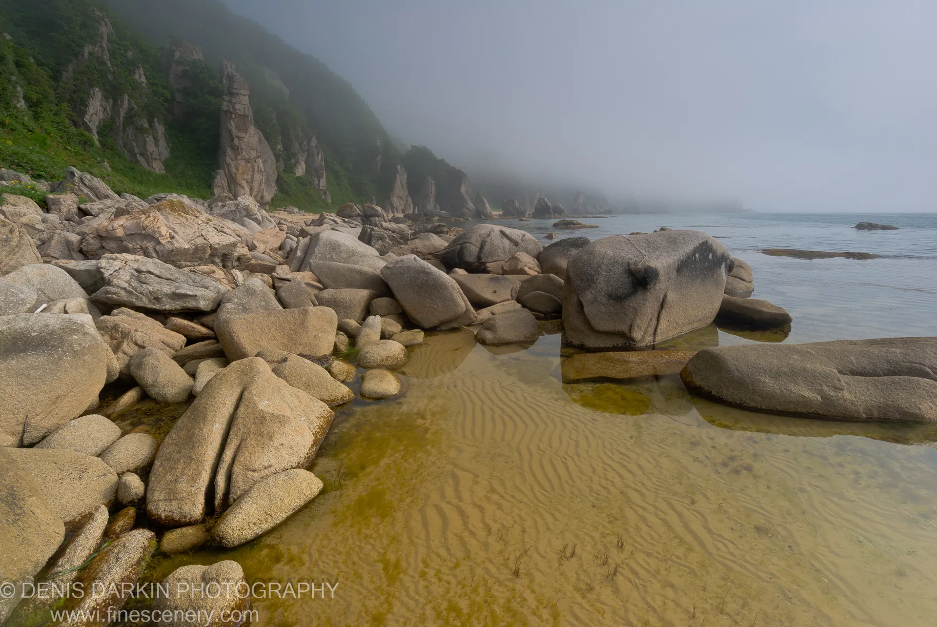 Weathered boulders rise from shallow water along the Lazovsky Reserve coast, with a steep green cliff and misty horizon beyond.