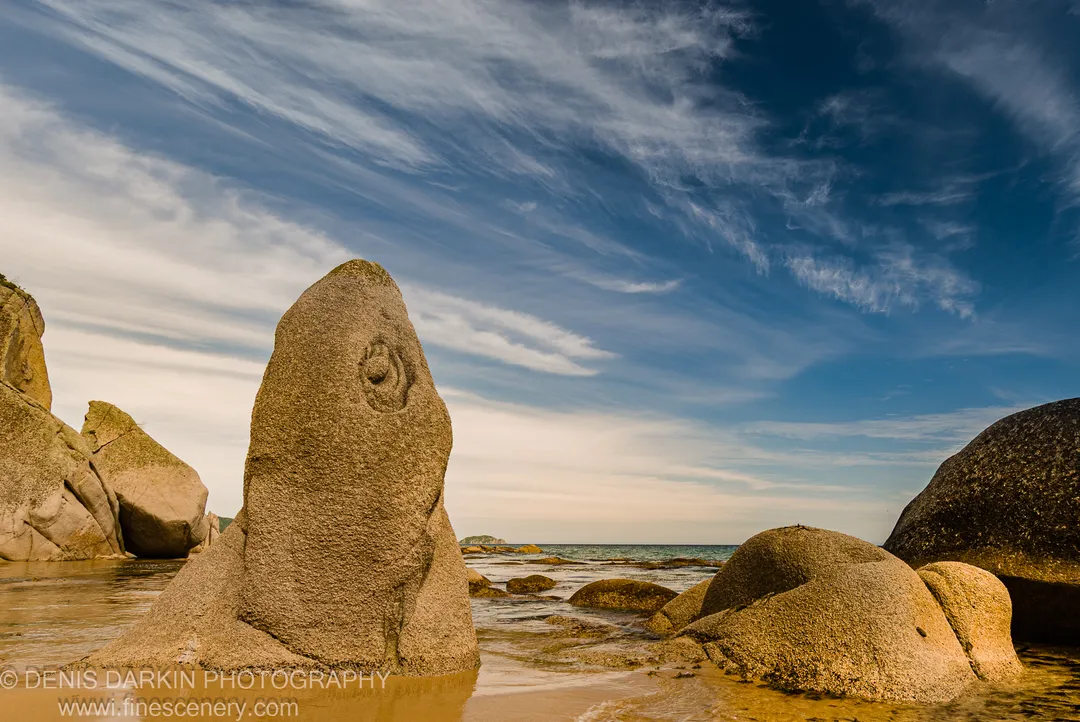 A spiral carving near the apex of a granite boulder at Tachingouza Bay, lit by afternoon sun against a blue sky with cirrus clouds.