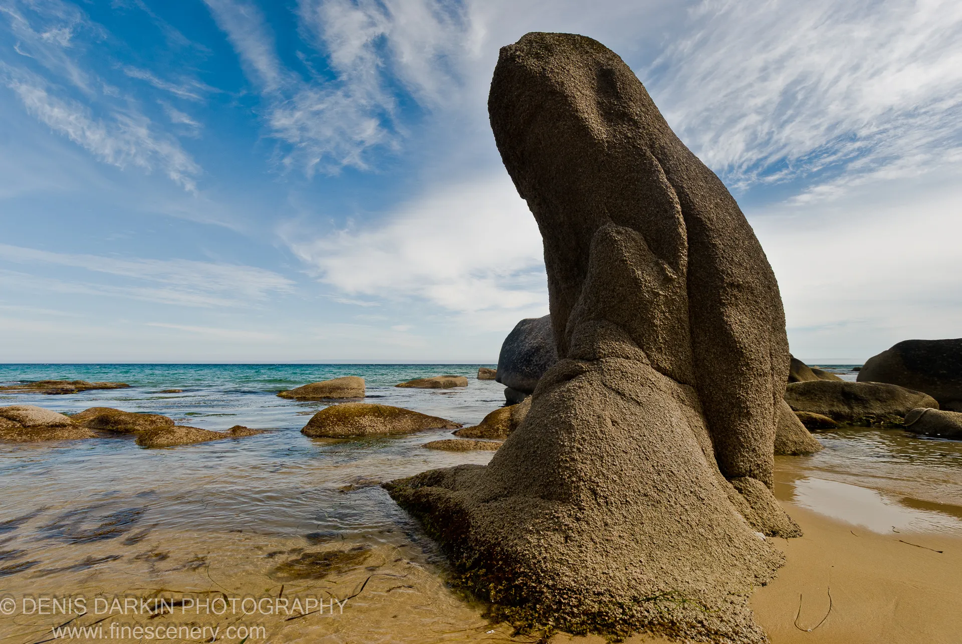 A massive vertical granite rock face rises from a sandy shore beside shallow water scattered with boulders, Tachingouza Bay.