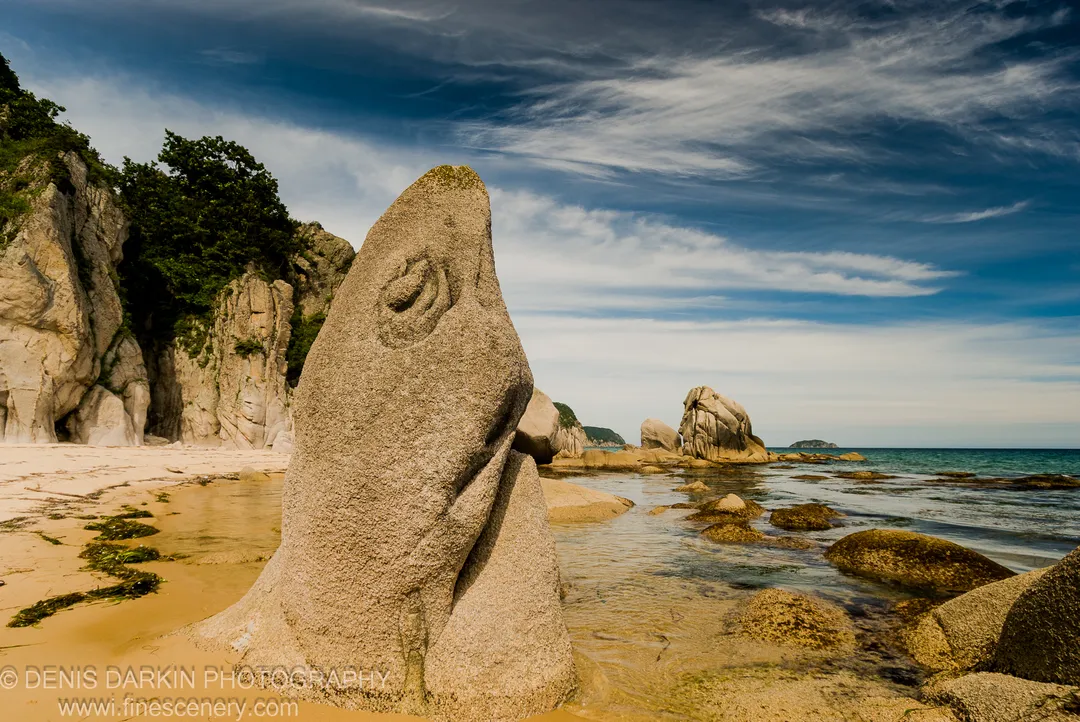 A large bulbous granite monolith stands above turquoise water, seaweed-strewn sand, and scattered boulders at the shore of Lazovsky Reserve.