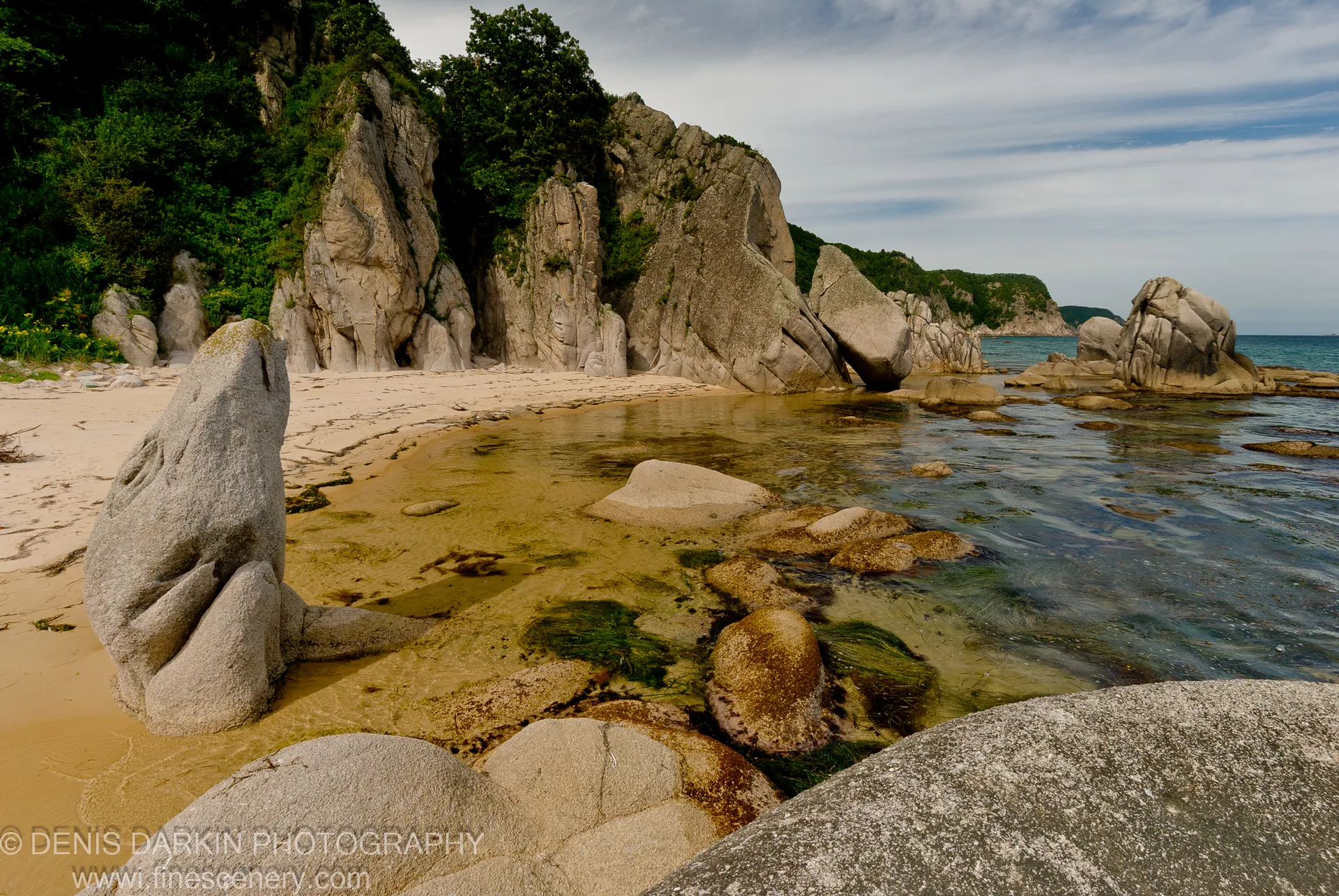 An upright stone stands in the surf at a sandy cove in Lazovsky Reserve, with lush vegetation on the cliffside above.