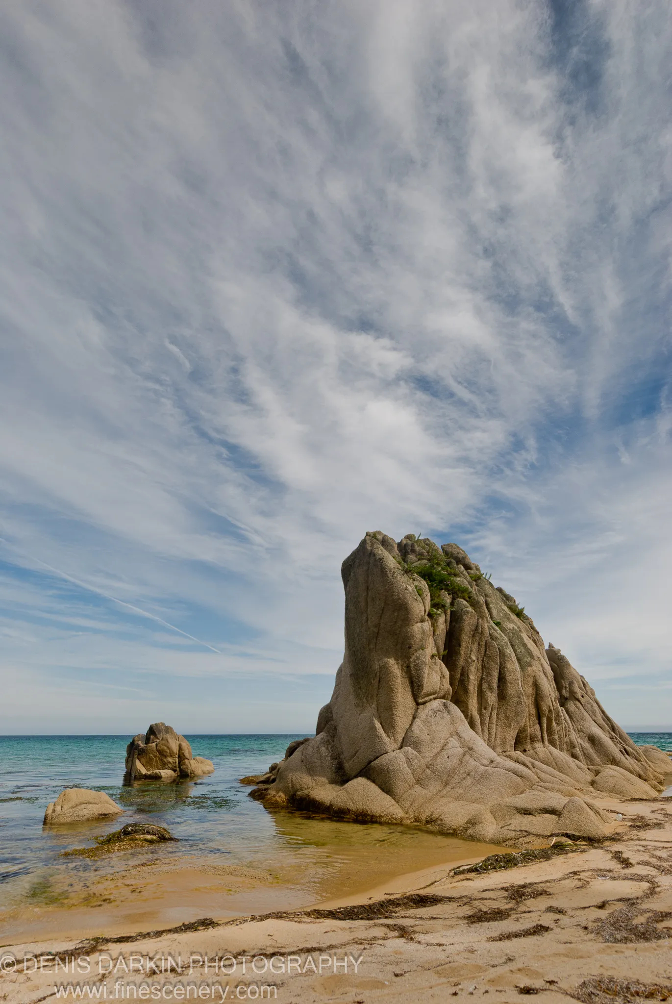 A massive rock formation rises above a sandy beach scattered with dark seaweed at Tachingouza Bay, with a smaller stone in clear water beyond.
