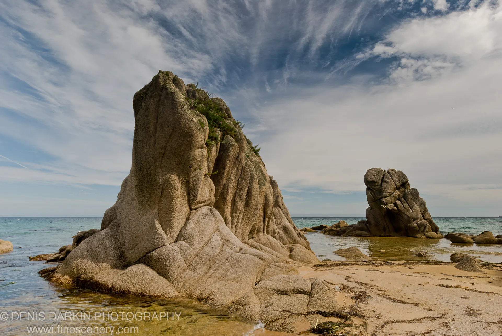 A tall granite monolith with deep vertical fissures stands in shallow water beside a smaller rock stack under cirrus-streaked sky, Lazovsky Reserve.