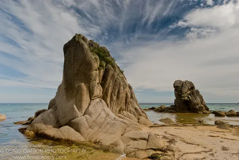 A tall granite monolith with deep vertical fissures stands in shallow water beside a smaller rock stack under cirrus-streaked sky, Lazovsky Reserve.