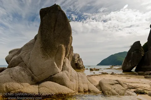 View through a natural arch between massive granite boulders at Tachingouza Bay, with a calm sea and distant green headland beyond.