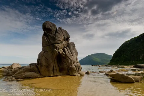 A granite monolith rises from amber tidal waters with soft reflections, set against lush hills and scattered clouds, Lazovsky Reserve.