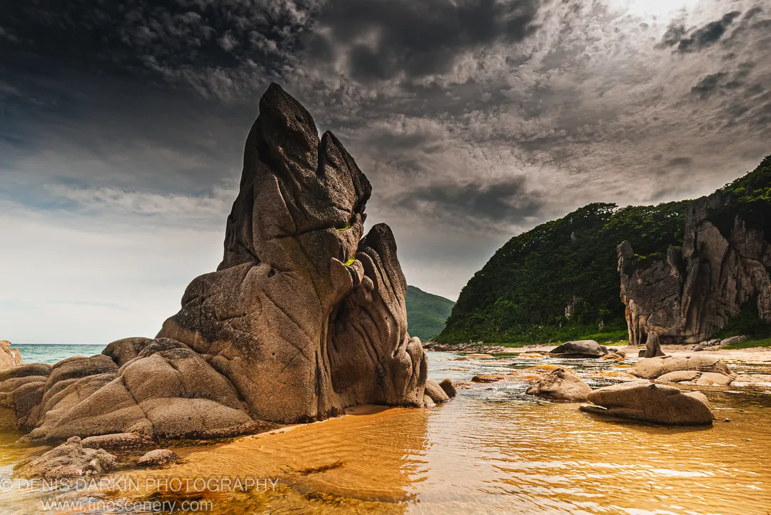 A towering granite rock with deep fissures at Tachingouza Bay, with a lush green hill descending to a sandy beach and dark clouds above.