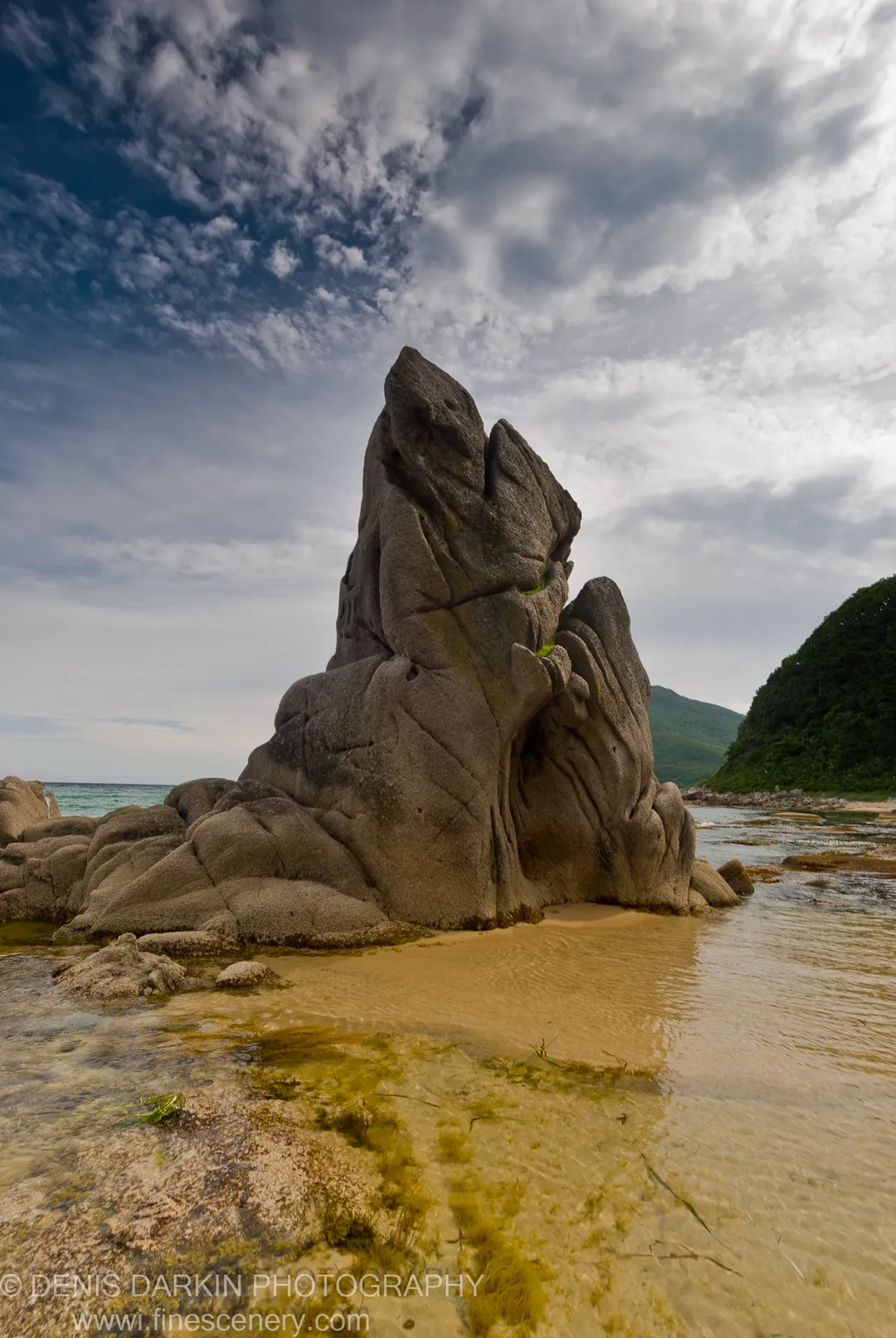 A tall granite rock formation rises from a sandy inlet, with shallow seaweed-patched water and a deep blue cloud-streaked sky, Lazovsky Reserve.