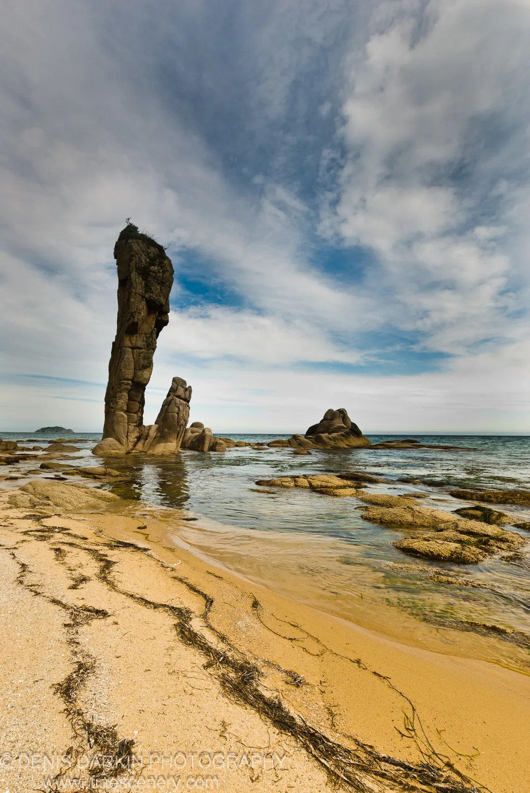 A tall sea stack above tangled kelp and golden sand, with a streaky cloud sky and calm water, Lazovsky Reserve.