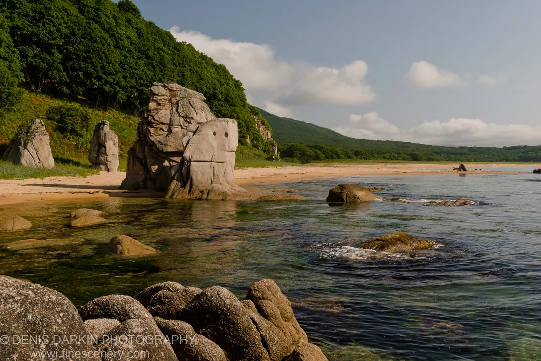 Large weathered granite formations on a sandy shore at Tachingouza Bay, with transparent water revealing submerged rocks and a forested coastline beyond.