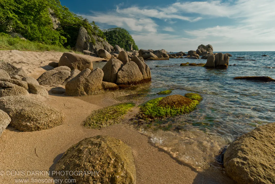 Moss-covered boulders rest on sand and in shallow water along Langou Bay, backed by forested hillsides.