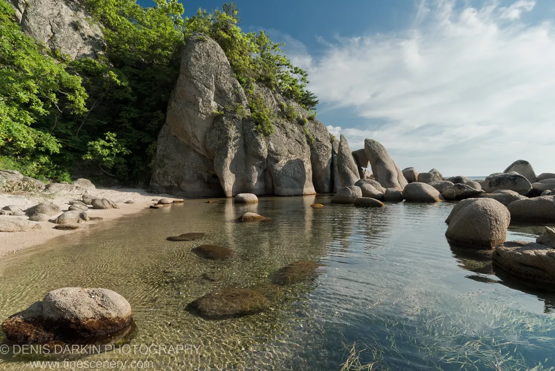 Clear water reveals submerged grasses and stones, with massive granite boulders and pale curved sand at Langou Bay.