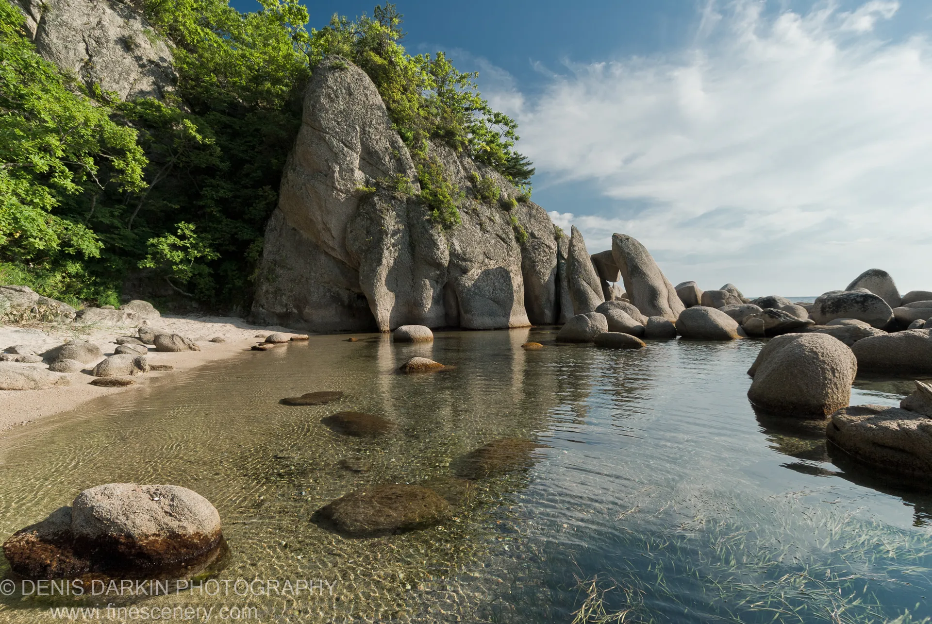 Clear water reveals submerged grasses and stones, with massive granite boulders and pale curved sand at Langou Bay.