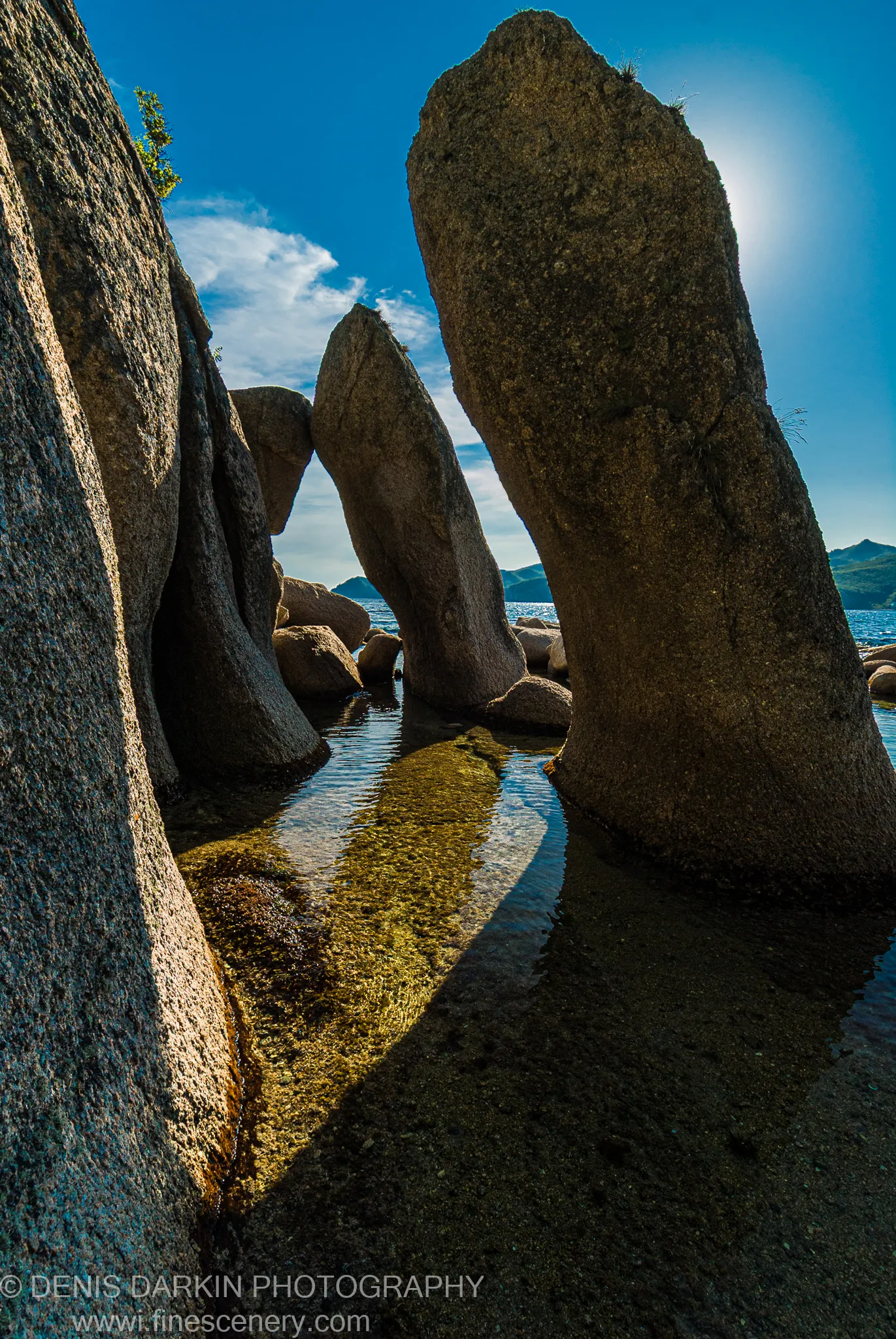 Two granite monoliths at Langou Bay form a natural arch above a tidal pool, their rough stone faces lit against a blue sky.