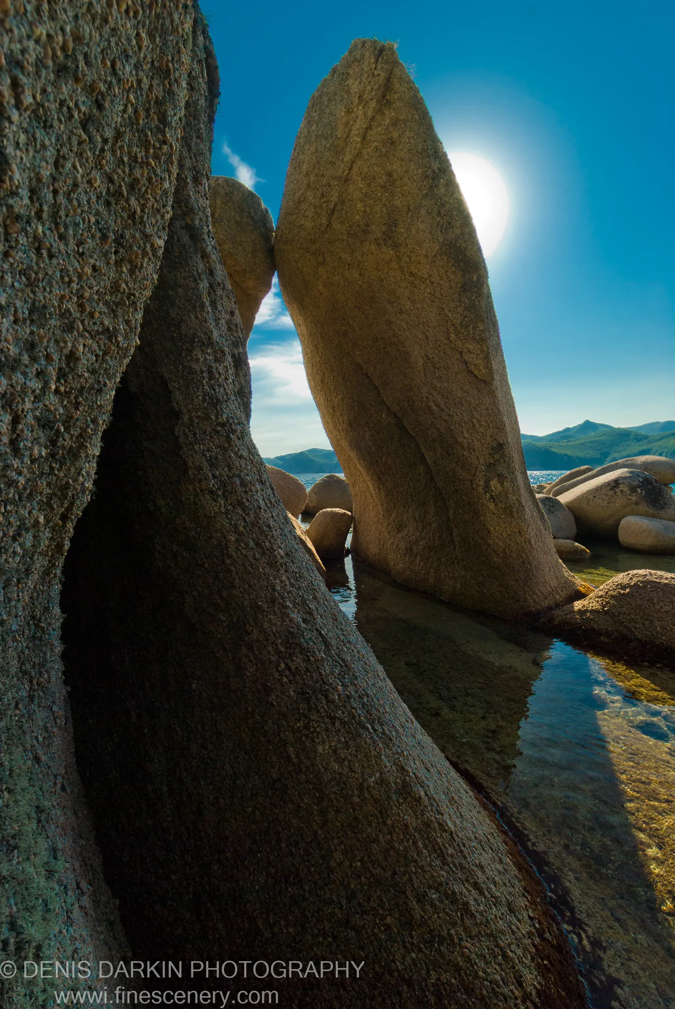 Granite boulders rise from clear shallow water at Langou Bay, backlit against the summer sky with deep shadows between the stone faces.