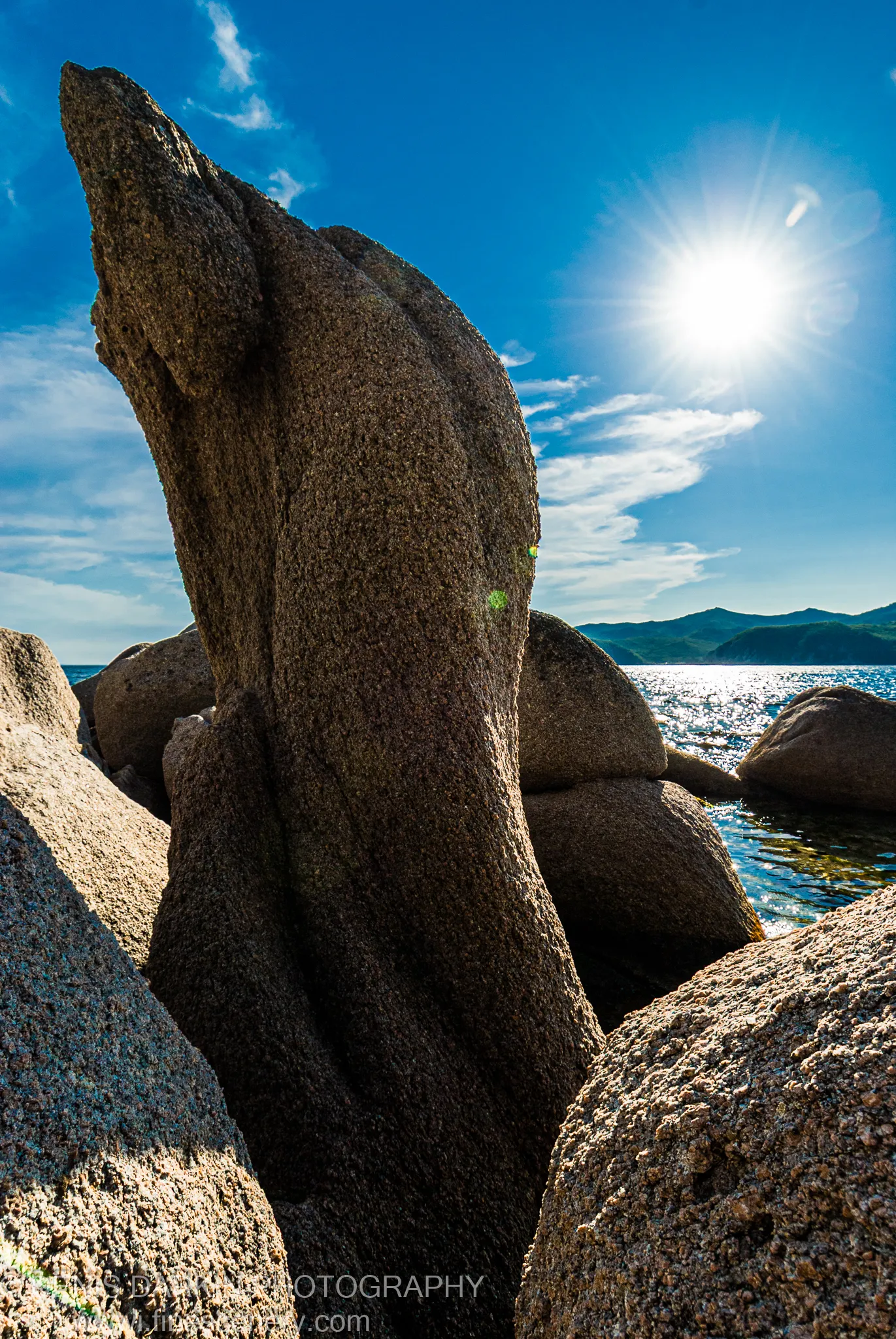 Sunlight forms a starburst above granite rock formations at Langou Bay, the sea surface below shimmering with reflections against a deep blue sky.
