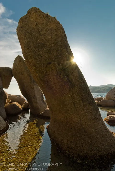 Tall granite pillars at Langou Bay rise from shallow water, sunlight bursting through a narrow gap between the stones.