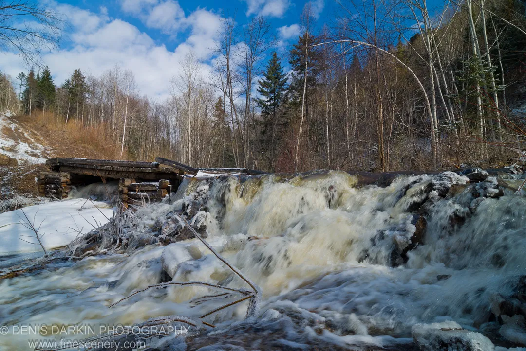 Rushing meltwater passes a log weir on the Vinogradovka in April, icy banks and bare trees below a deep blue midday sky.