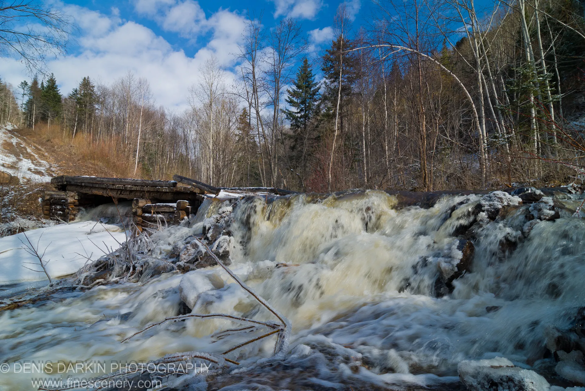 Rushing meltwater passes a log weir on the Vinogradovka in April, icy banks and bare trees below a deep blue midday sky.